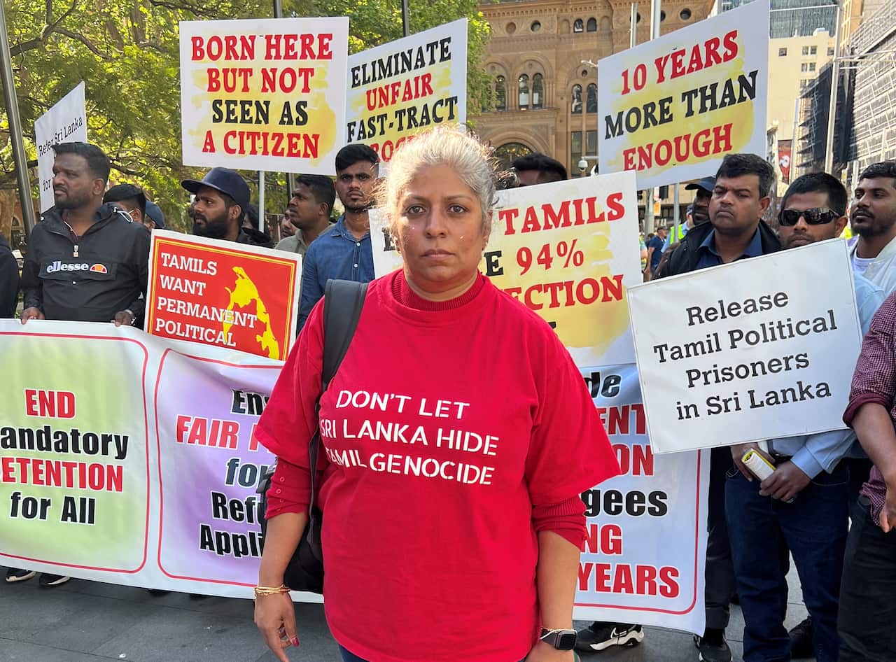 A woman at standing in front of people holding signs. She wears a red T-shirt which reads: 'Don't let Sri Lanka hide Tamil genocide'.