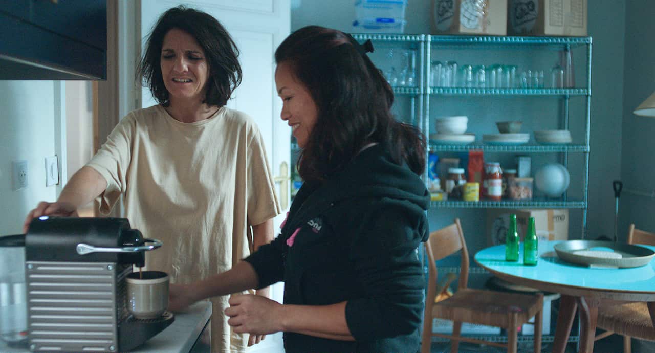 Two women, one of them looking a little dishevelled, stand in a kitchen beside a coffee machine, as it pours coffee into a cup.