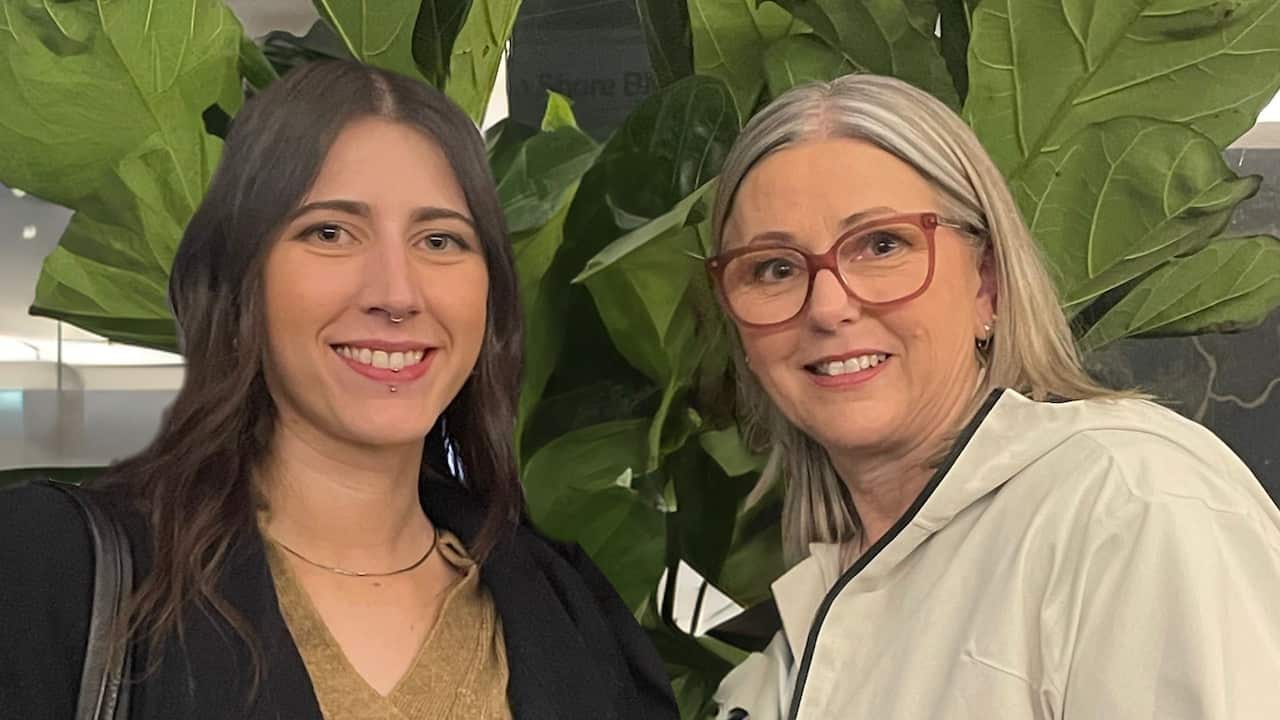 a woman in her 20s with dark hair, septum and lip piercings smiles with a middle aged woman with grey hair and wide rimmed glasses in front of a plant