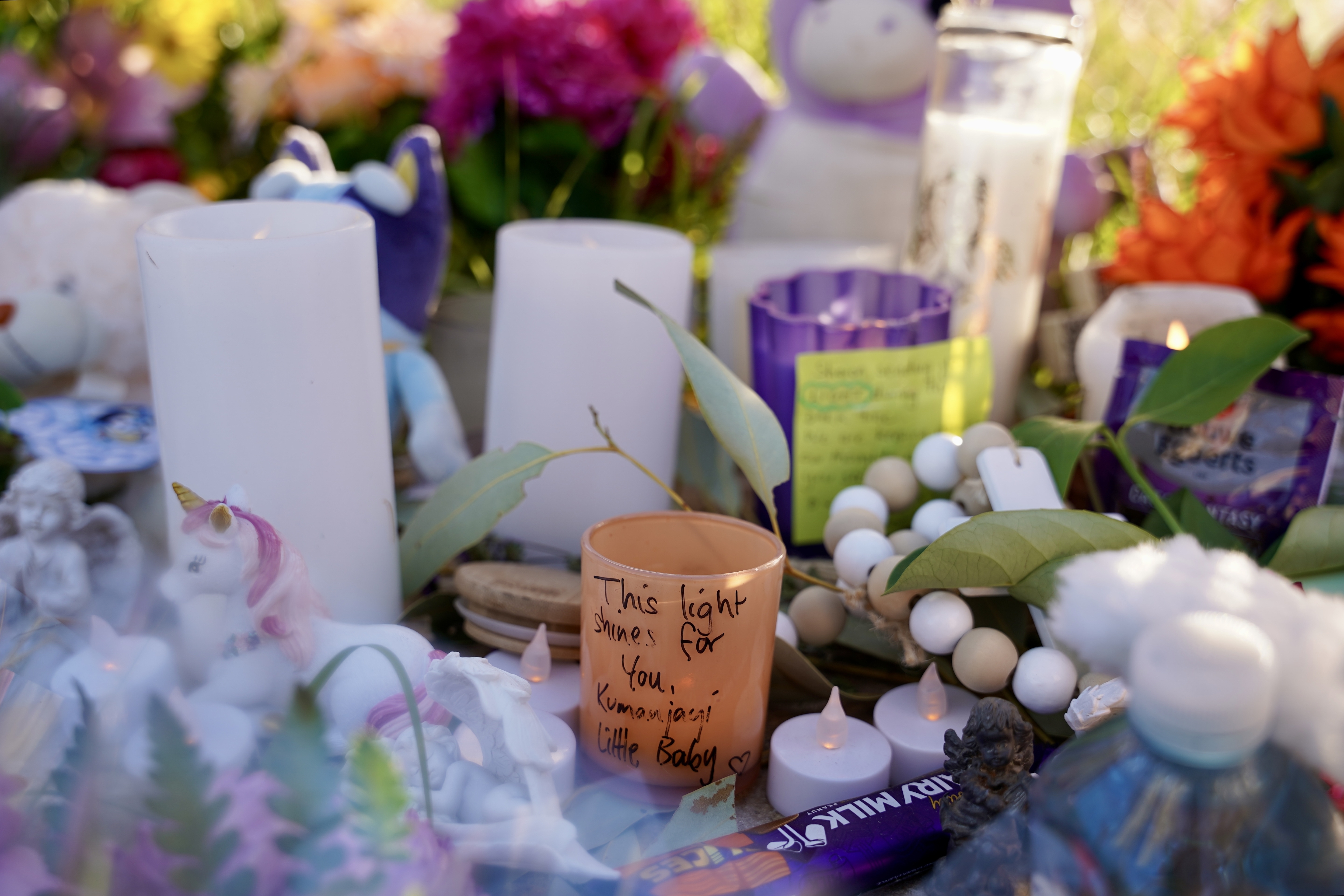 A cluster of tributes including candles, eucalyptus leaves, and teddy bears. A candle in the foreground has writing on it that reads: "This light shines for you, Kumanjayi Little Baby".