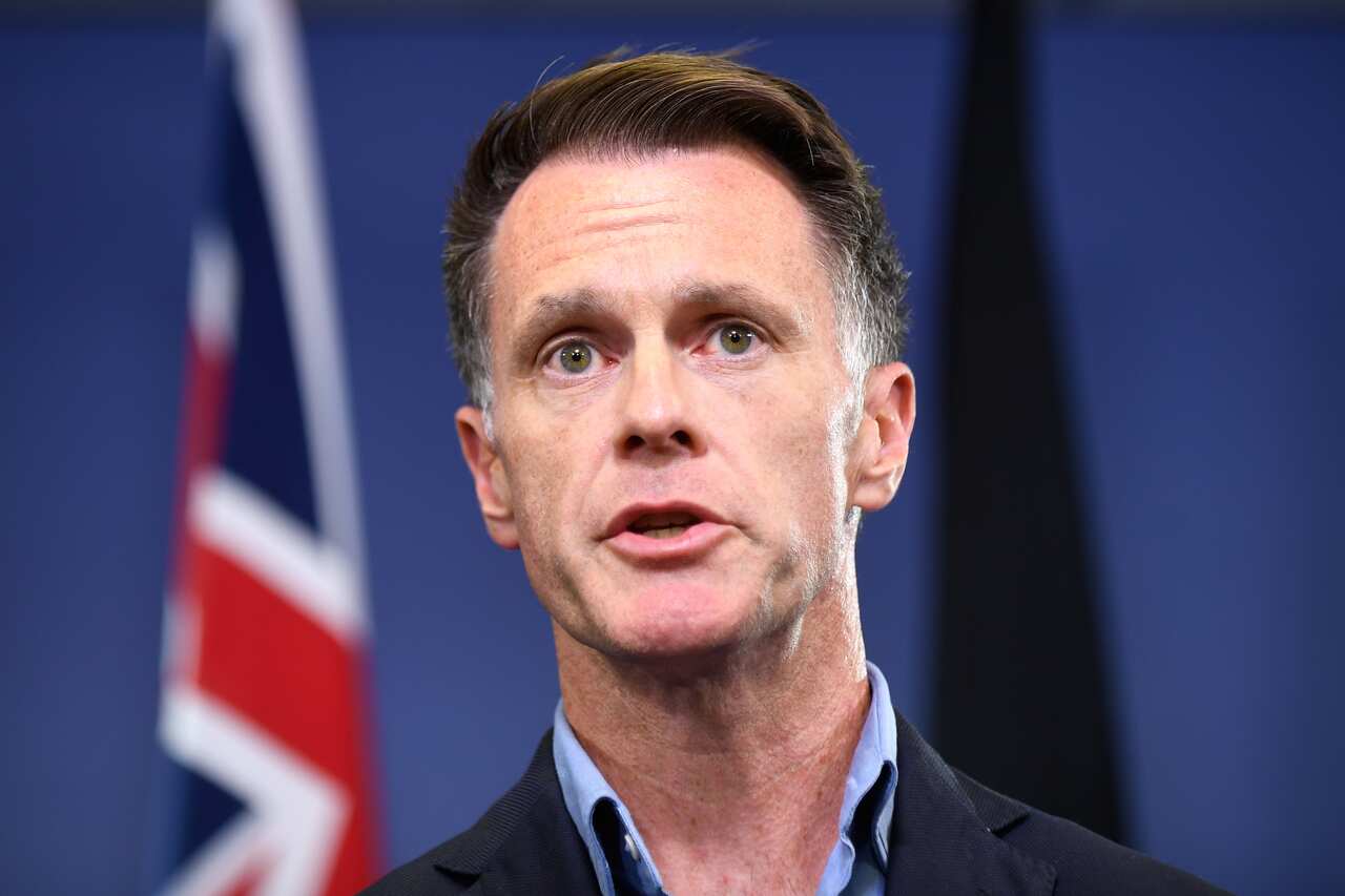 A man in a dark suit stands in front of a background with an Australian flag visible