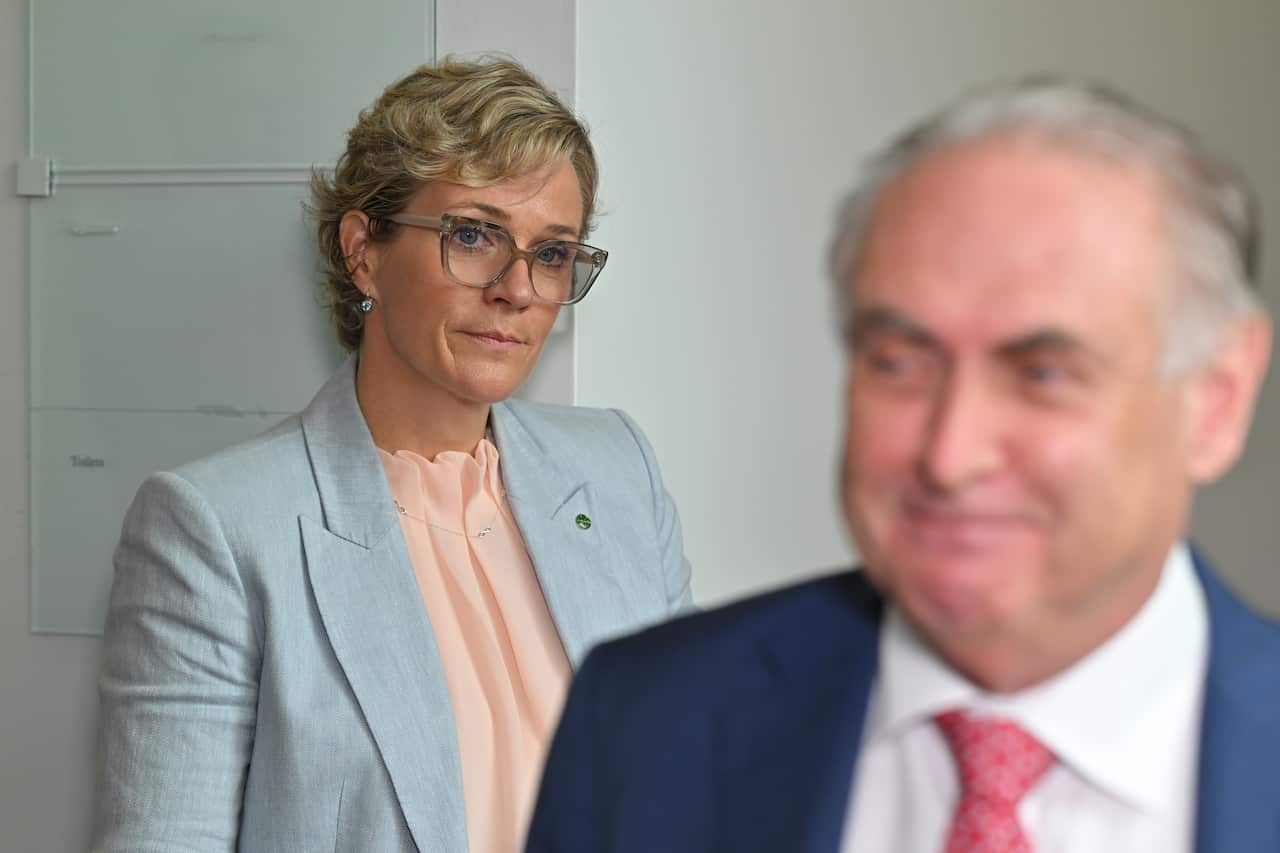 A woman in glasses and a blazer looks ahead at a man in a blue suit.