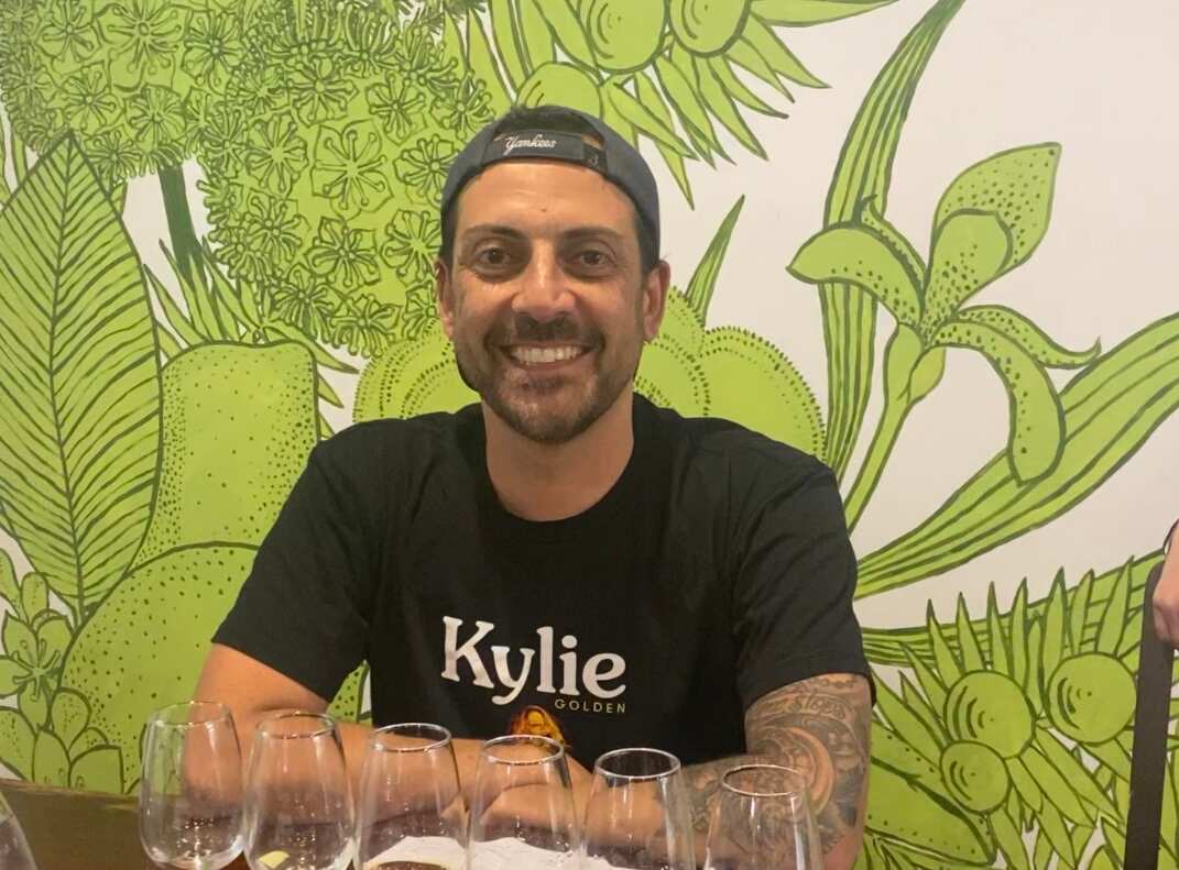A man wearing a black t-shirt featuring the word Kylie in while letters smiles as he sits in front of a row of empty glasses.