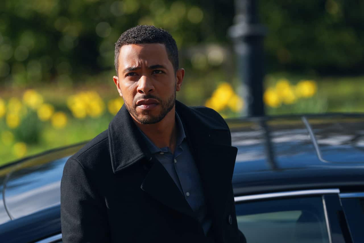 A tense looking man in a dark coat looks to be on the move, with a car just visible behind him, and a garden out of focus behind car. 