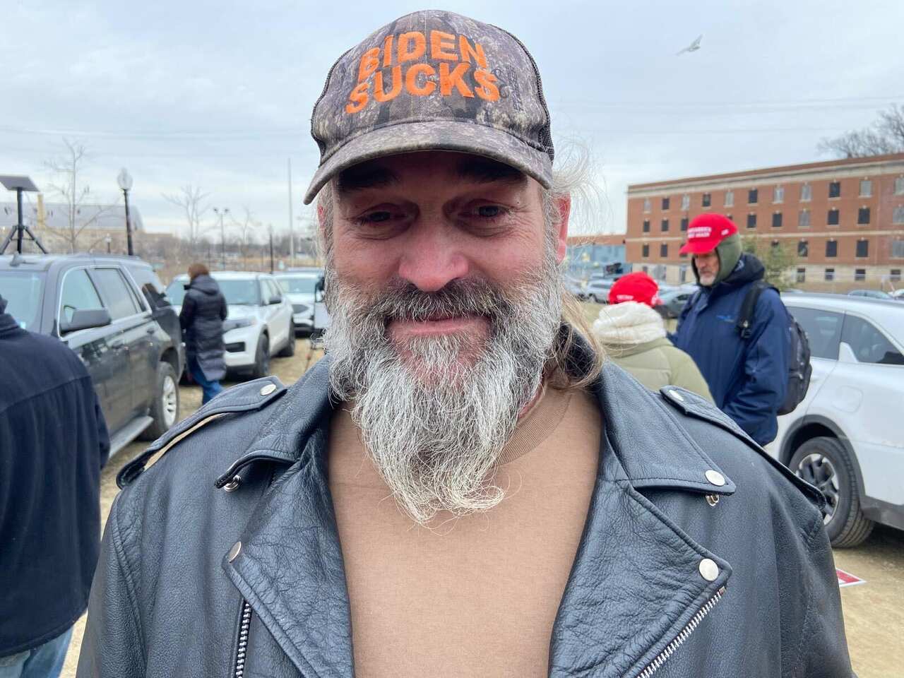 A man with a beard wearing a 'BIDEN SUCKS' hat and a black leather jacket.