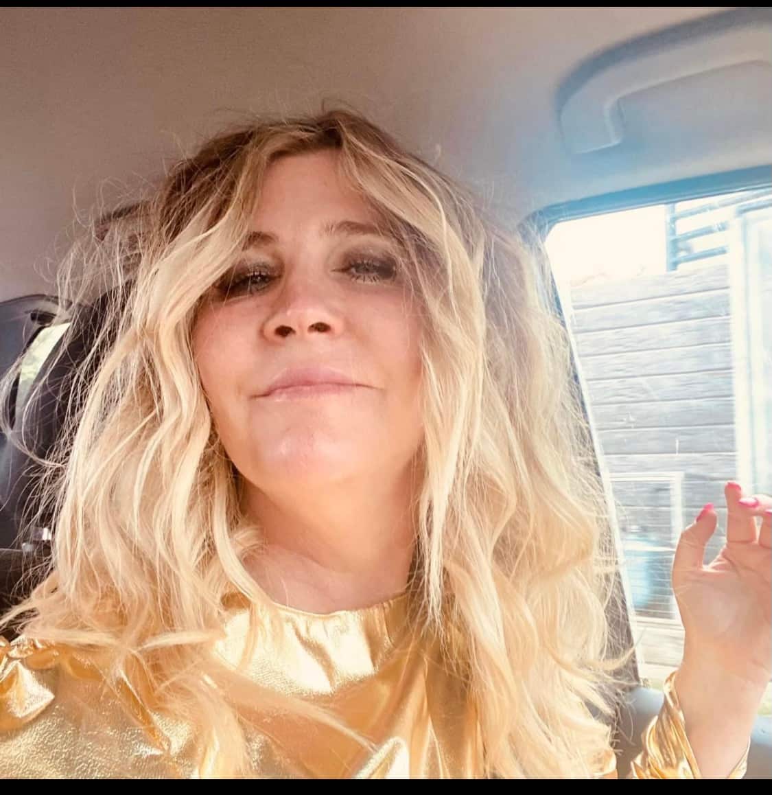 A woman with long blonde hair sitting in a car smiles at the camera.