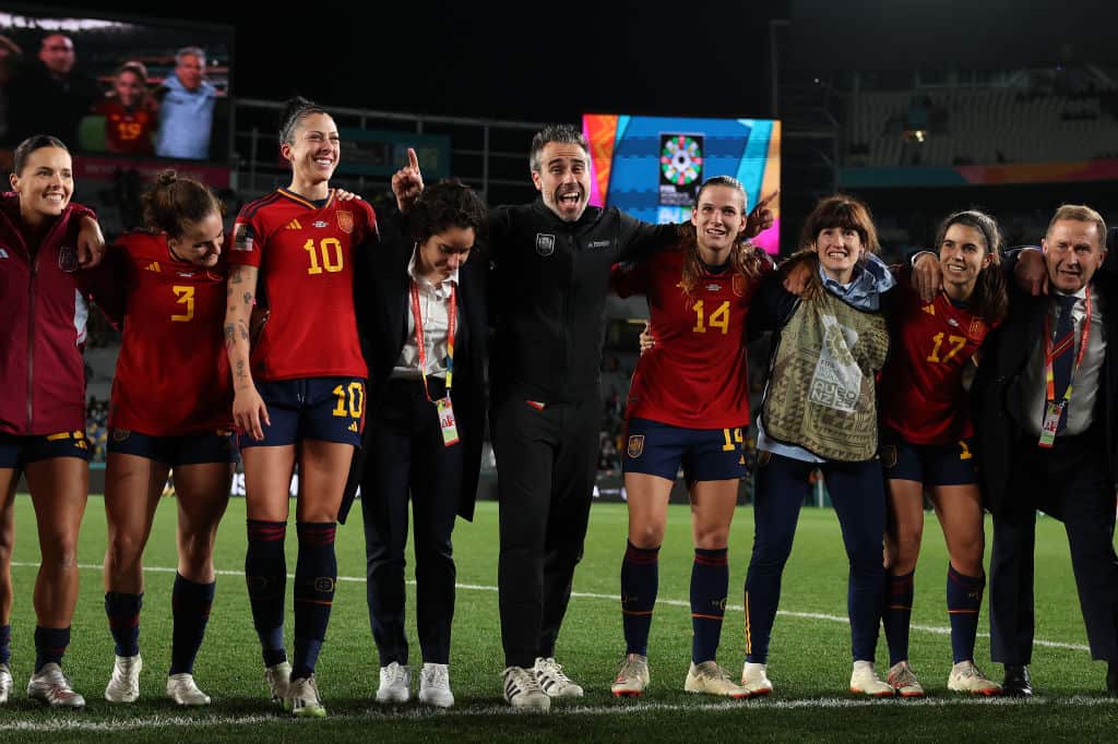 A man with a grey beard and wearing black stands on a field in a line of female footballers wearing red jerseys. There is a also a woman wearing black standing next to him and a man in suit at the right end of the line.