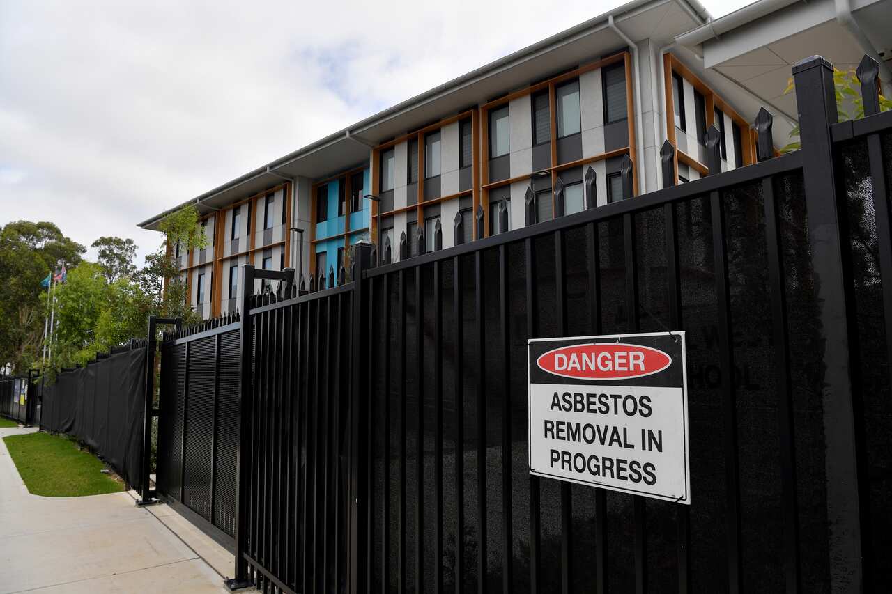 The school building is visible behind a fence covered in black plastic with a warning sign that reads: Danger! Asbestos removal in progress.