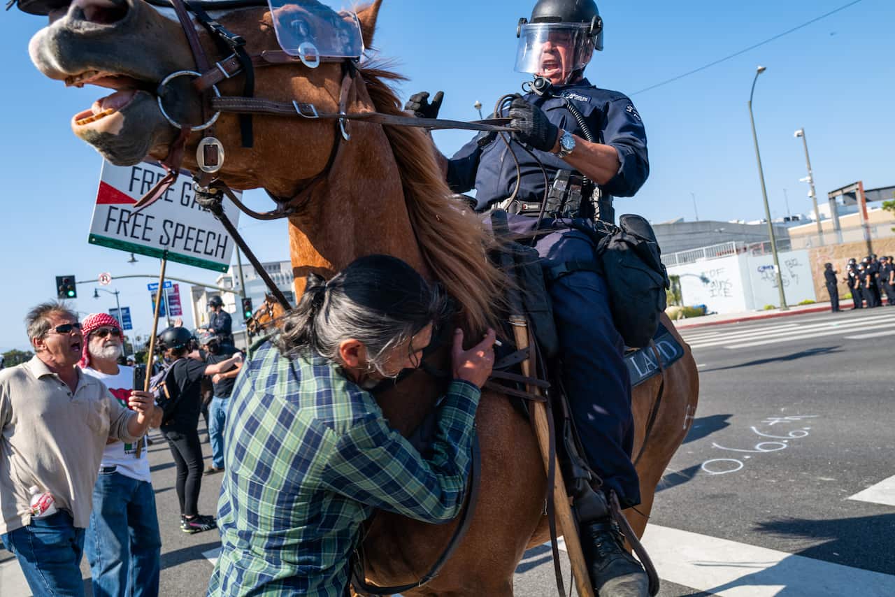A person in a green and blue shirt colliding with a horse being ridden by a police officer.