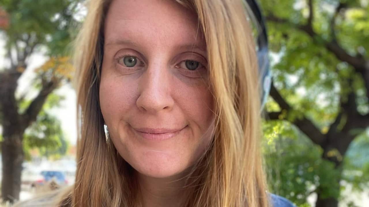 a blonde, blue-eyed young woman close up selfie in a park with blurred green leaves in the background
