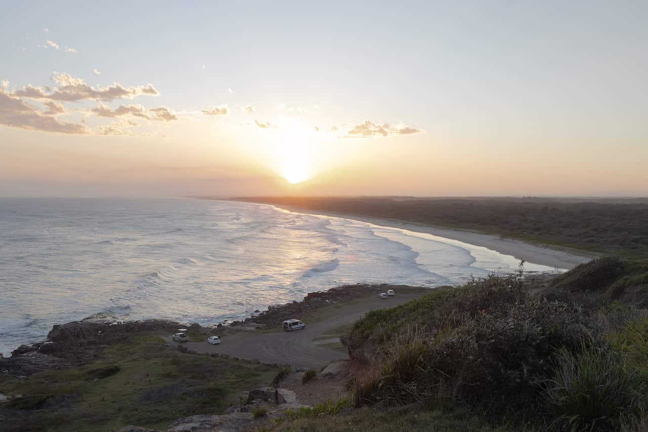 A coastal landscape view at sunset, showing waves crashing on a long beach backed by coastal vegetation, with a few cars parked near the water on the left.