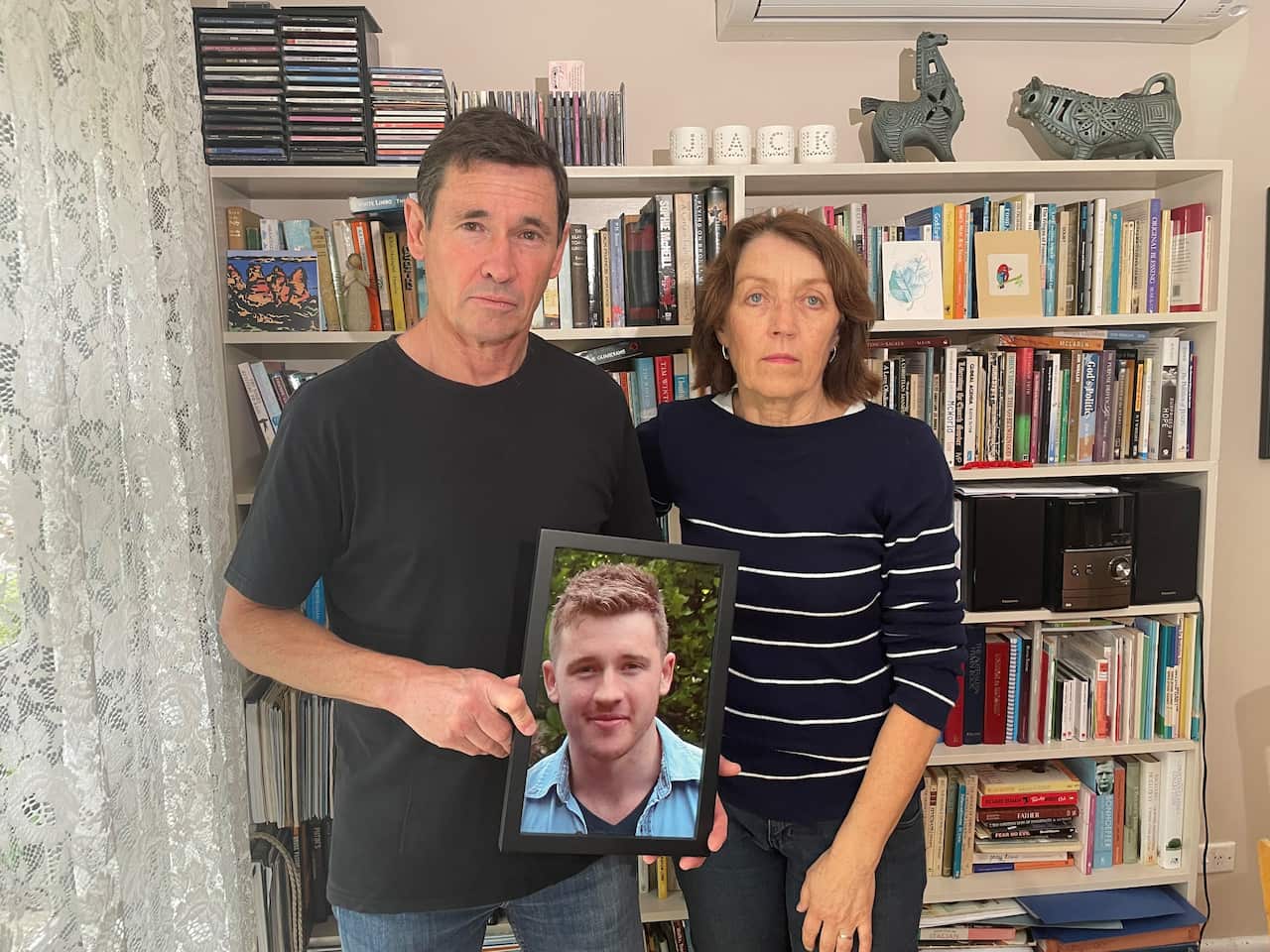A man and a woman standing in front of a bookcase and holding a framed photograh 
