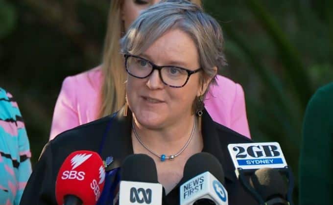 A woman with black-rimmed glasses stands in front of several microphones with News Outlet logos on them