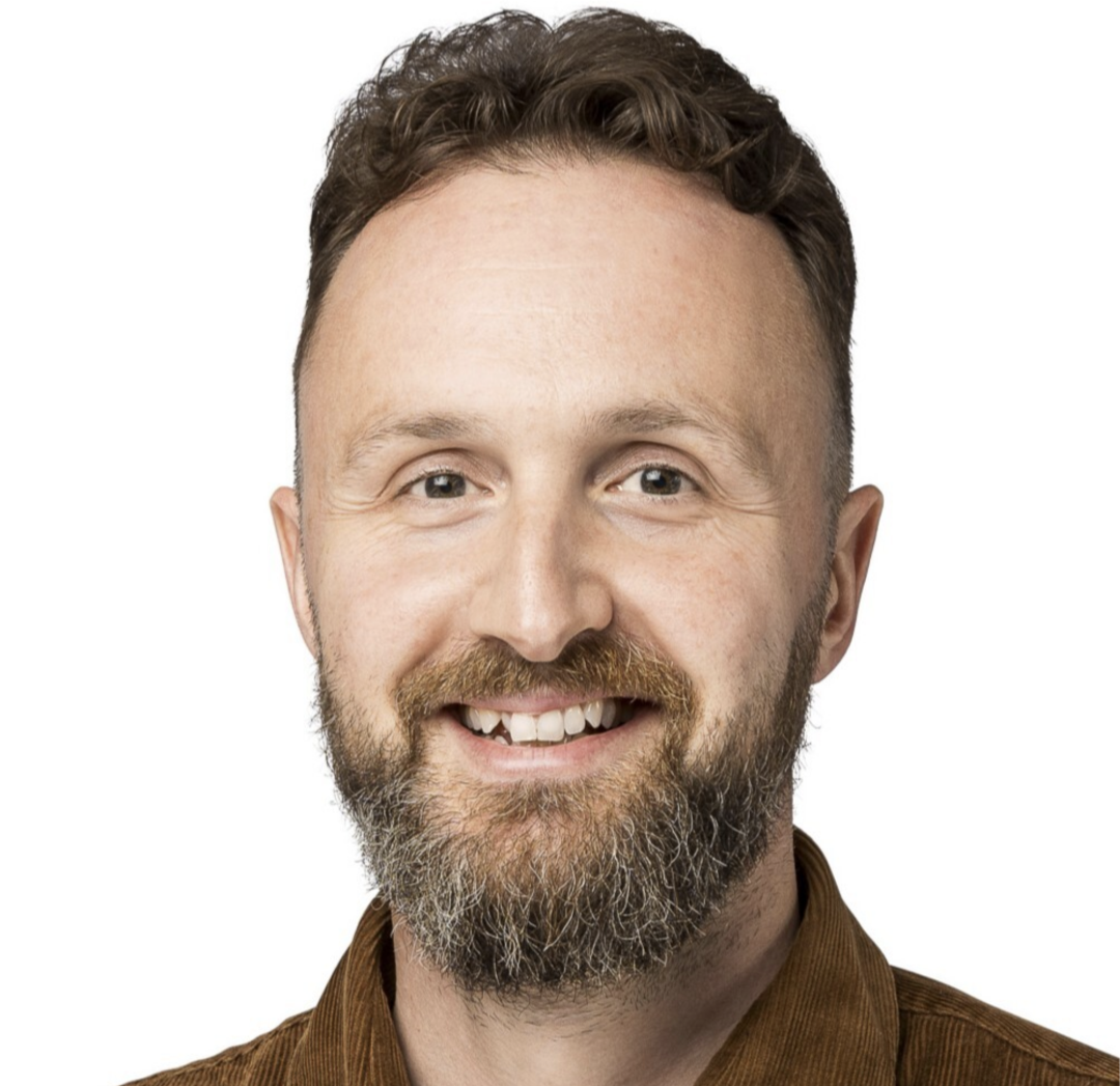 A man with a beard and a brown shirt smiles at the camera.