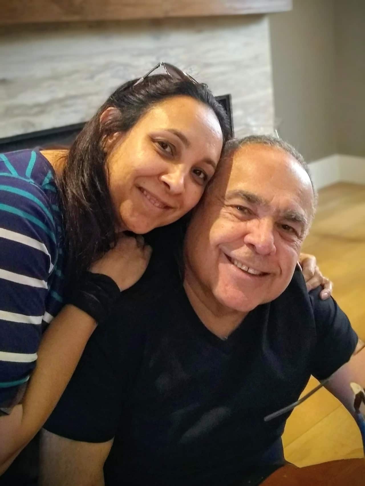 A portrait taken indoors of a smiling woman wearing a striped top with her arms around a smiling older man wearing a black top.