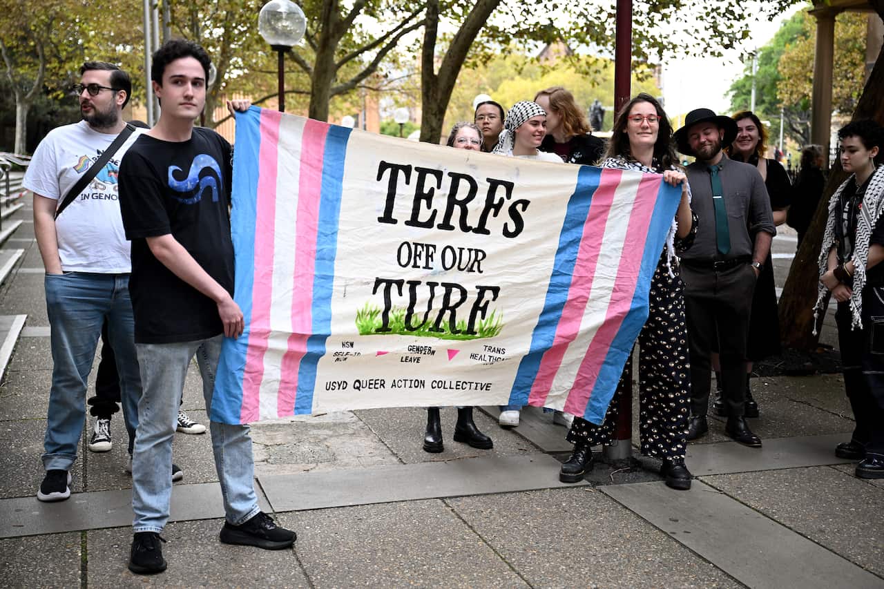 Protesters hold a sign with the pink, white and blue transgender flag colours and text reading "TERFs off our turf".