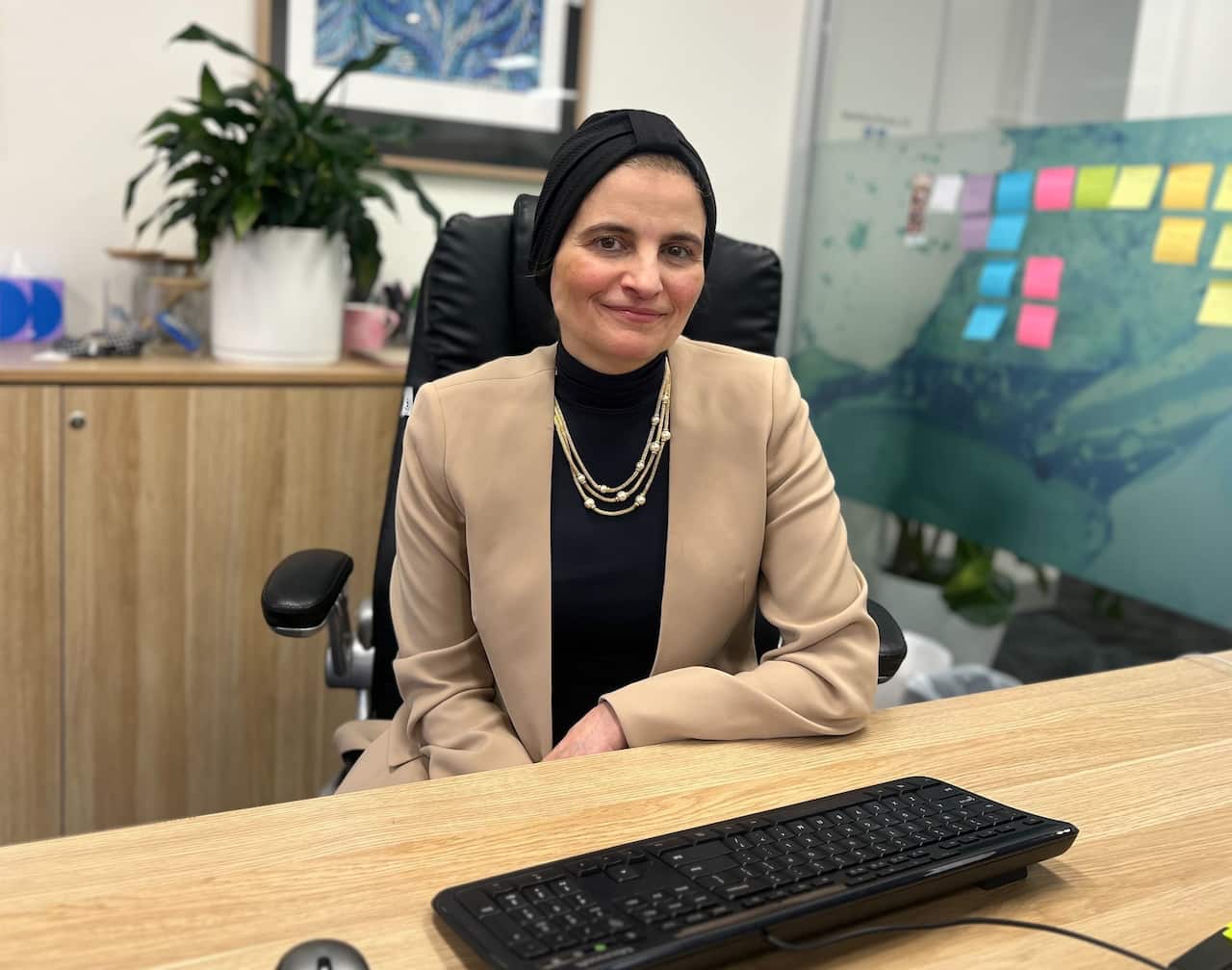 A woman in a beige blazer sits behind a desk, with a black keyboard in front of her. There is a plant and several colourful post-it notes in the background.