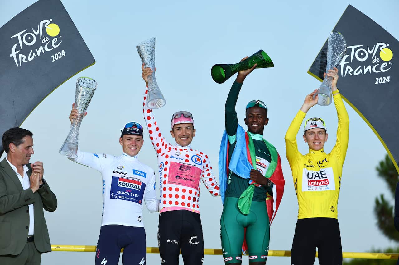 Four men in different-coloured cycling kits, including a white jersey, polka-dot jersey, green jersey, and yellow jersey, stand on a podium, raising trophies, with a man in a suit standing next to them and clapping.
