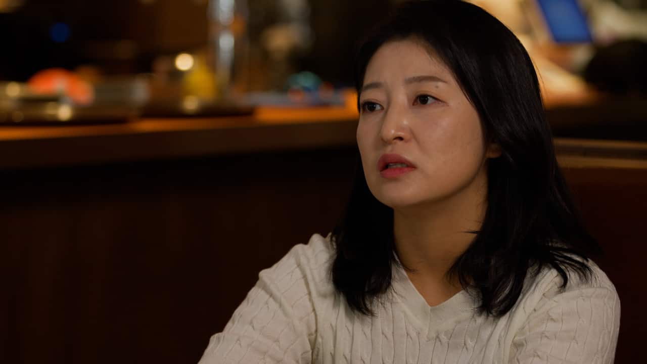 A woman in a white top sits in a cafe with a neutral expression on her face.