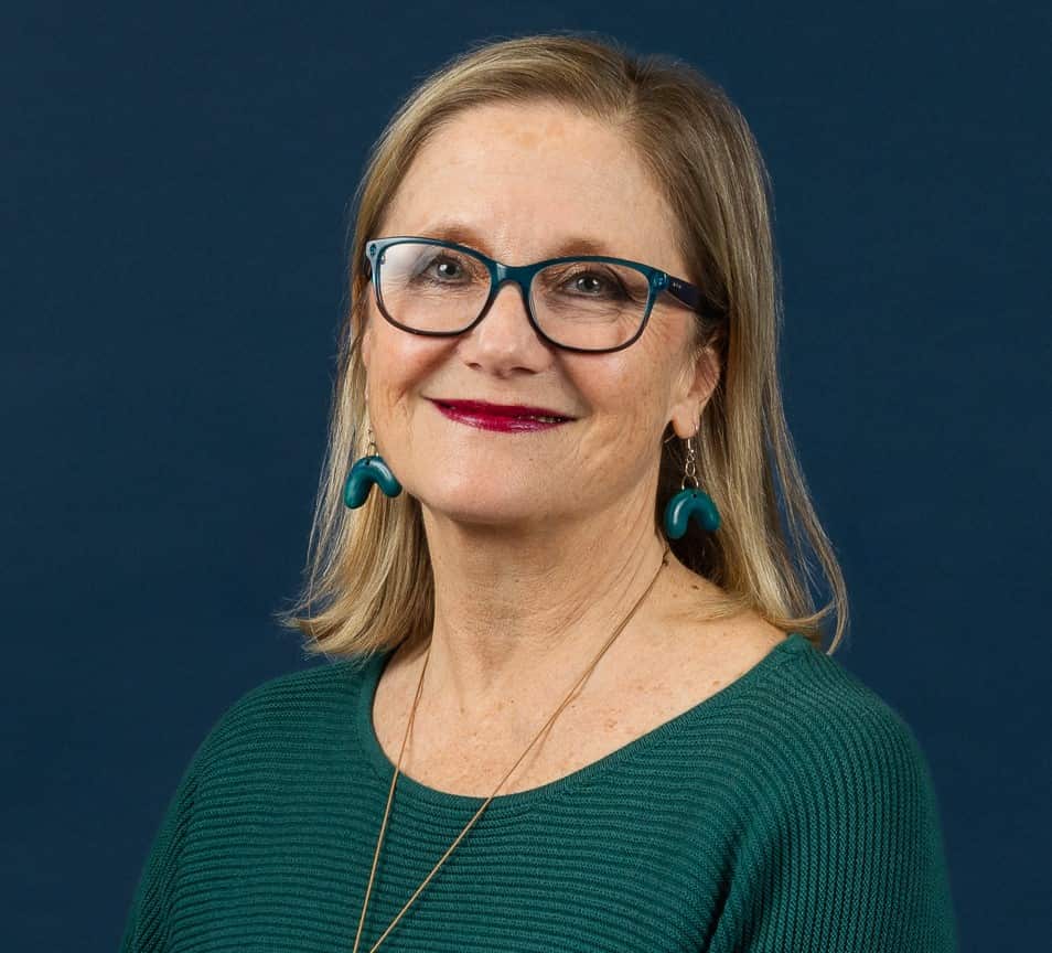 A headshot of a woman wearing a green top, glasses and green dangly earrings. 