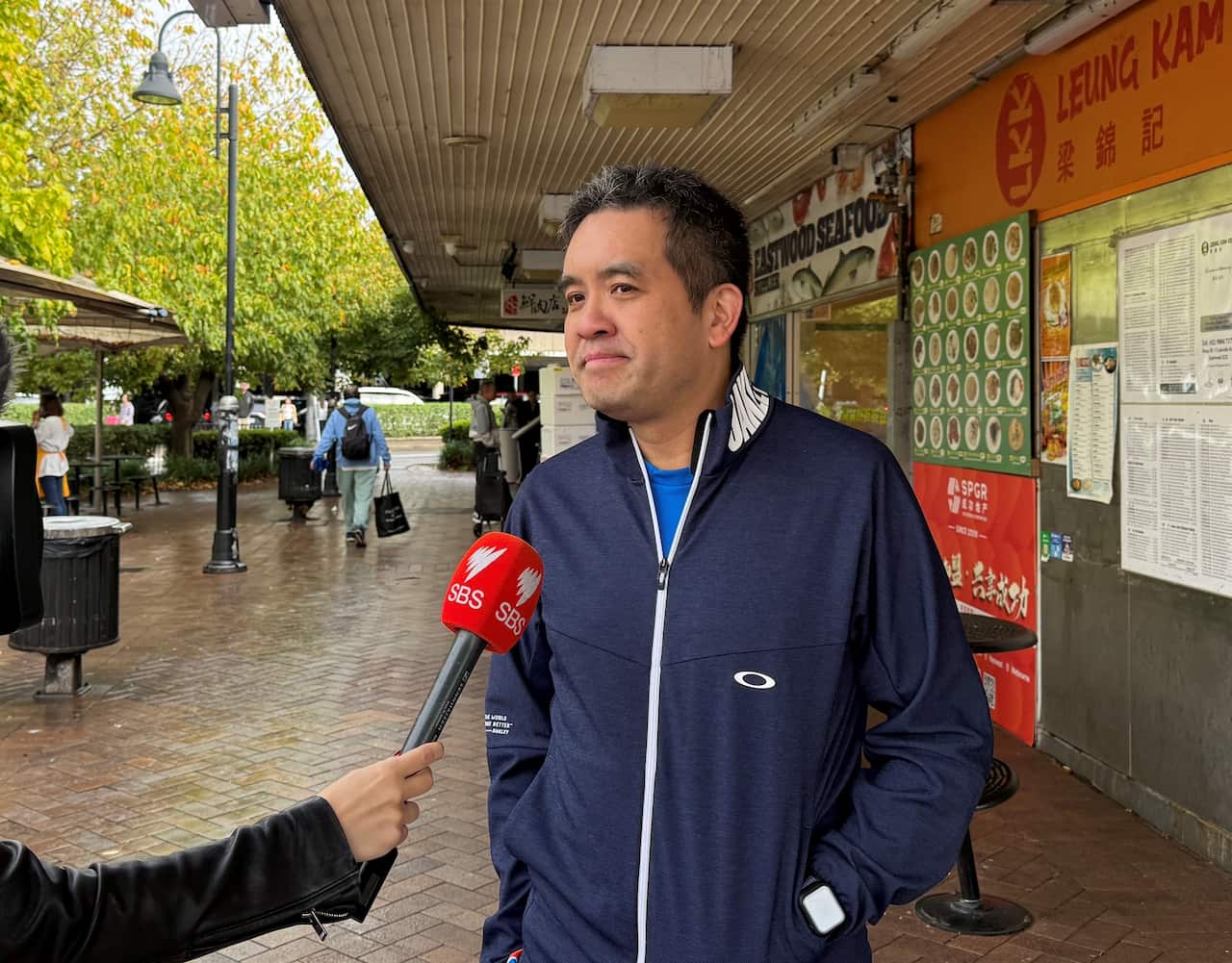 A man in a navy matching tracksuit, with a red microphone held up to his face.