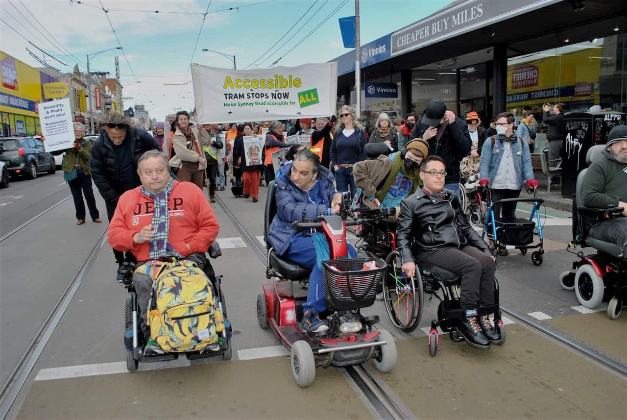 Protestors, some walking and others in mobility aids, make their way along the tram lines on Sydney Road, Melbourne.