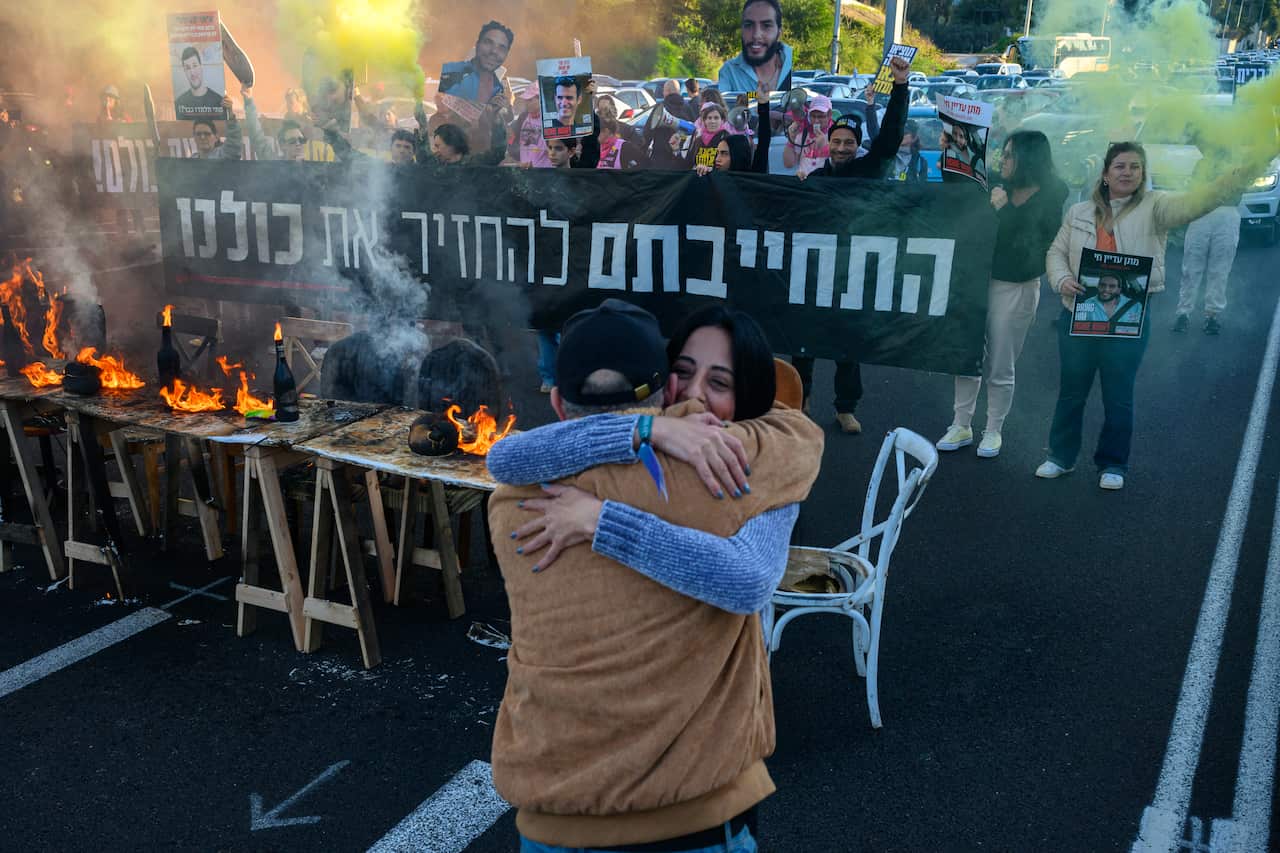 Two people embrace in the foreground while protesters behind them hold a large Hebrew banner and posters of hostages, framed by yellow smoke and small fires burning on a makeshift table.