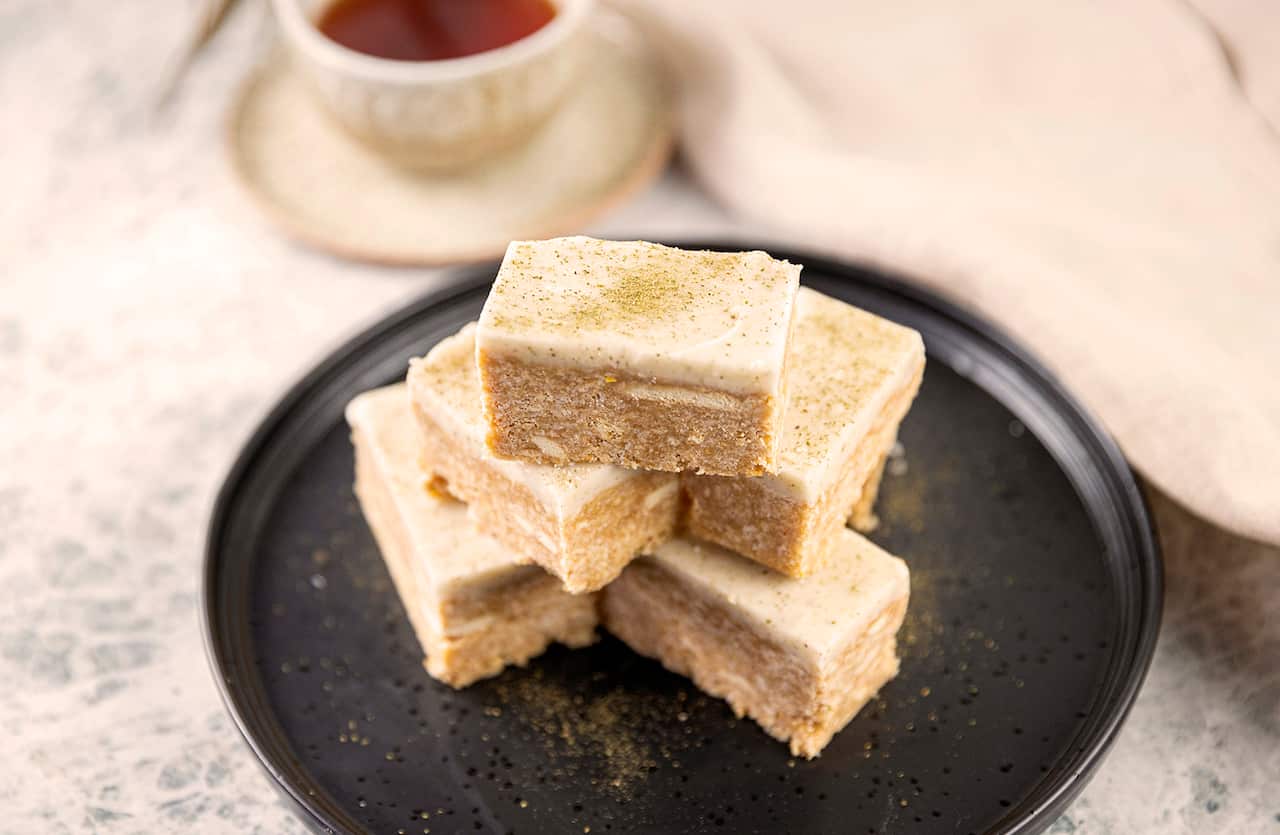 A three-deep stack of slice pieces sits of a flat black plate. Each piece of light brownish-caramel coloured slice is toped with pale icing.  