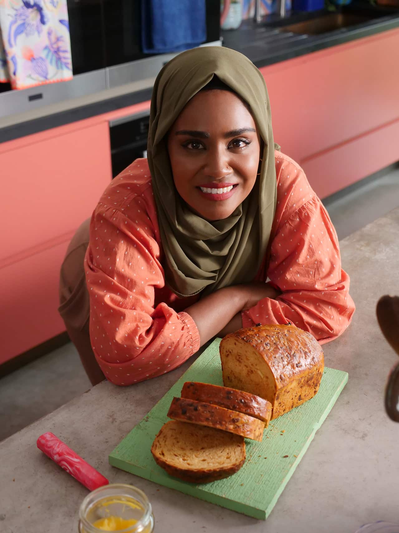 A woman in an orange top and olive green headscarf leands on a kitchen bench. A loaf of bread sits on a green board in front of her. A few slices have been cut from the loaf and sit, leaning, beside the rest of the loaf. 