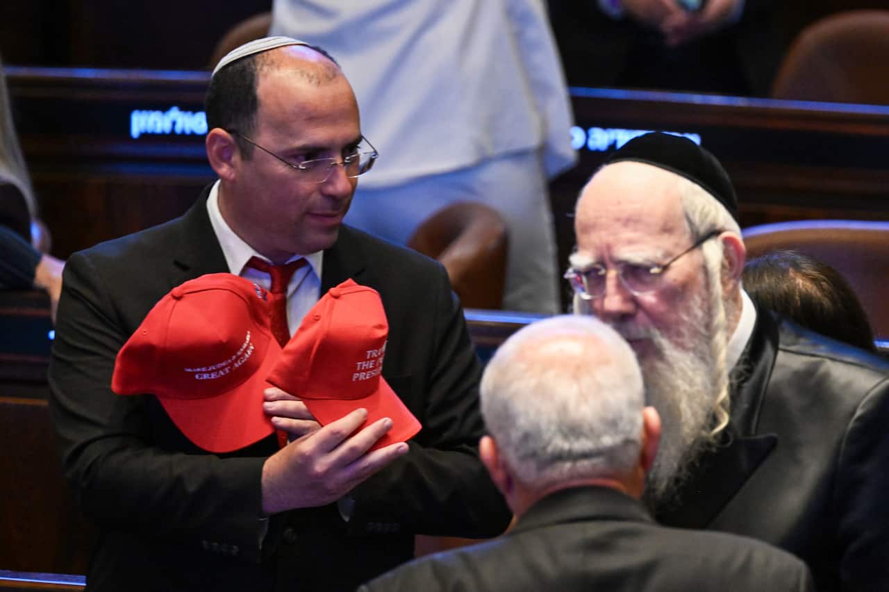 A man wearing a kippah holds two red caps. There are two elderly men standing next to him.