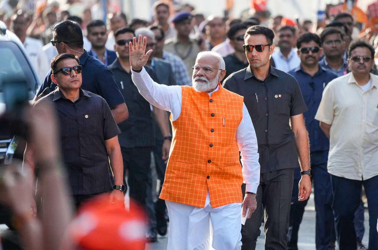 Narendra Modi, wearing an orange vest and white shirt and pants, walks in front of a crowd. He is waving with his right hand.