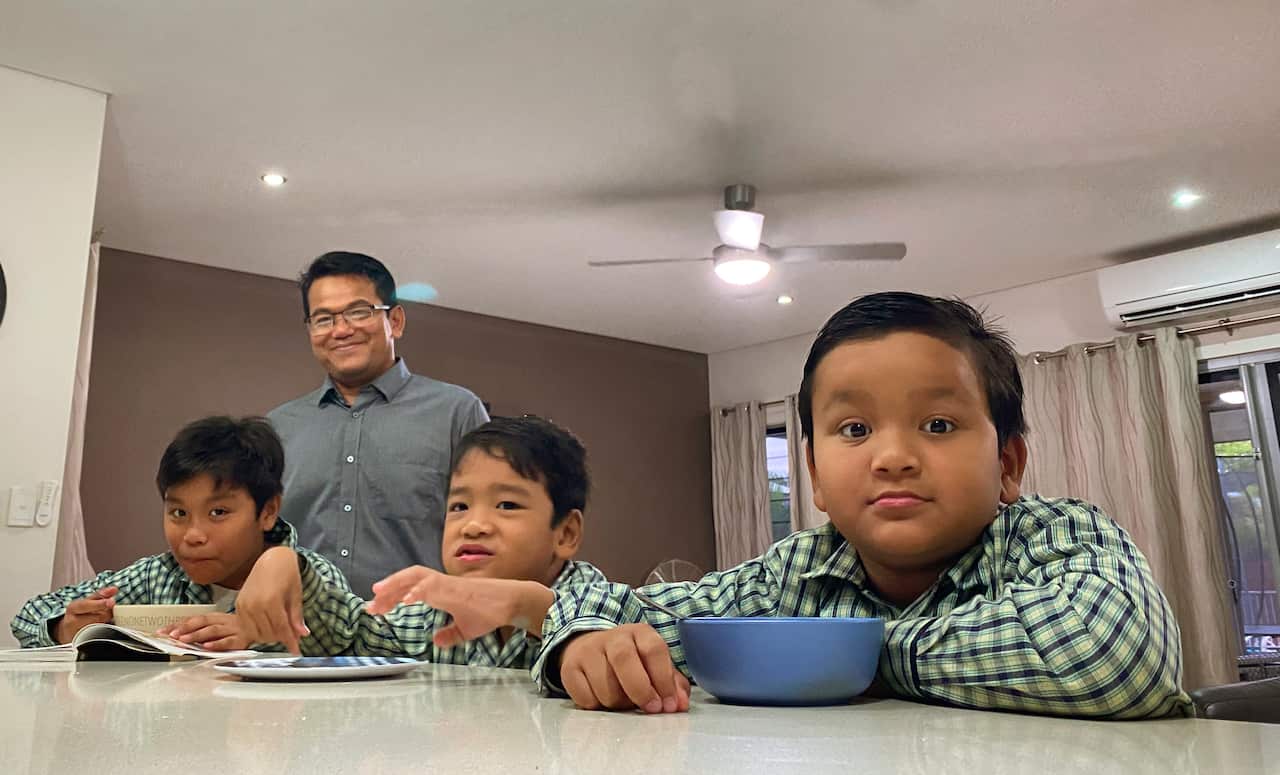 A man stands behind three children sitting at a kitchen bench.