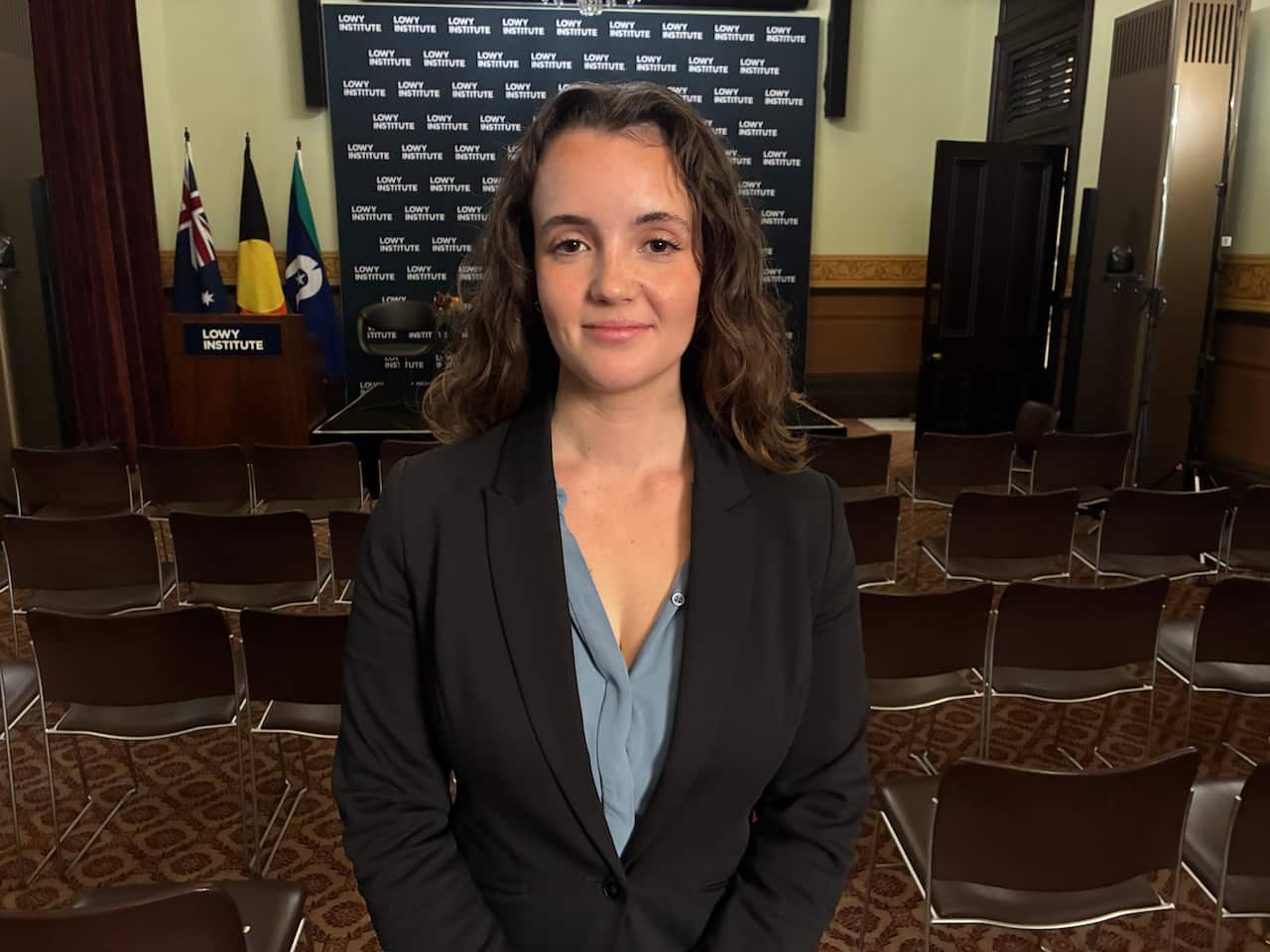 A woman in a dark blazer poses for a photo in front of rows of empty chairs.