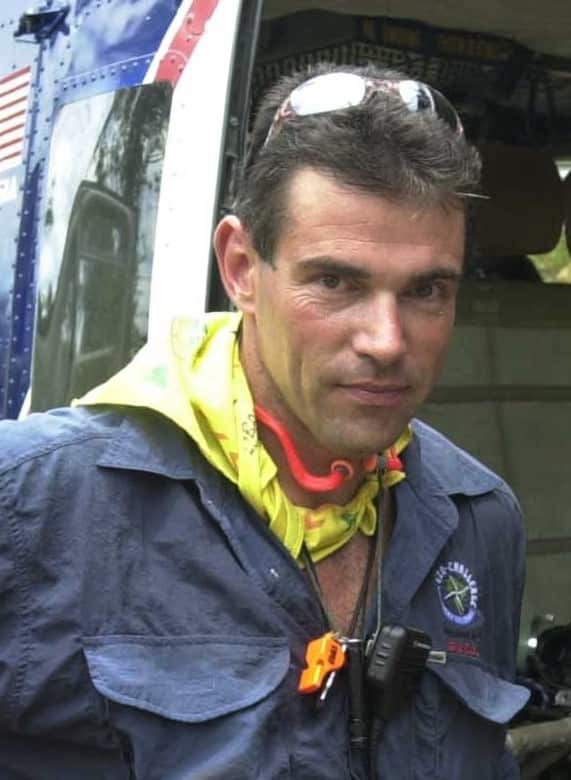A man in a blue shirt with a yellow scarf around his neck stands next to a helicopter doorway.