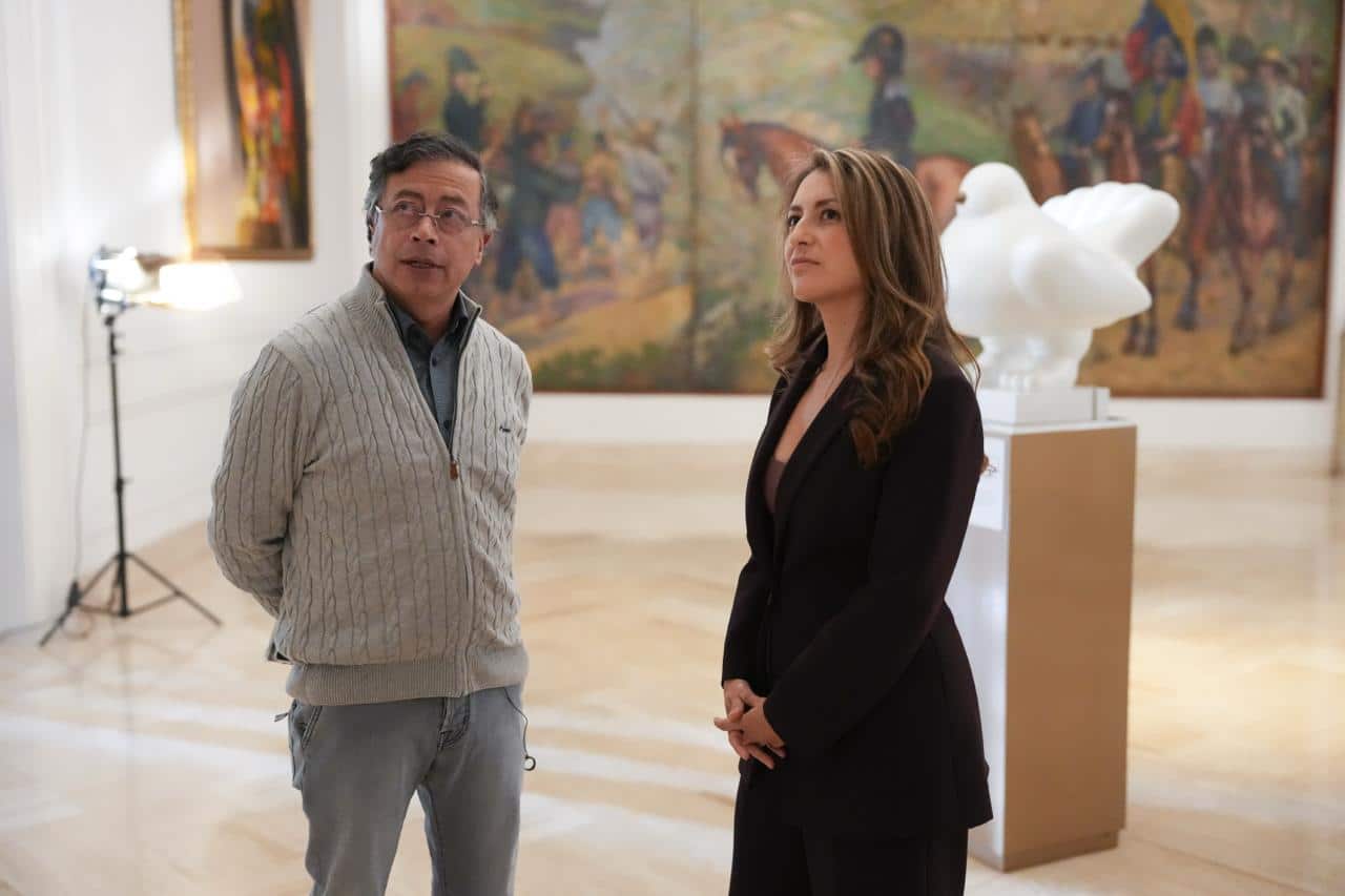 A grey haired man wearing glasses and a grey zip-up jumper stands next to a woman in a suit. They are looking at sometheing out of frame. They are standing in a large room, with paintings and a statue behind them, as well as a standing light used in TV recordings.
