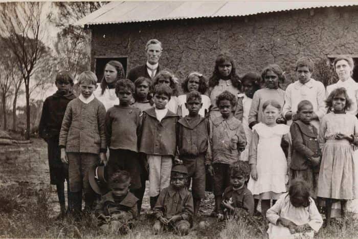 black and white photo of aboriginal children standing outside a simple building, with one white man standing at the back. 