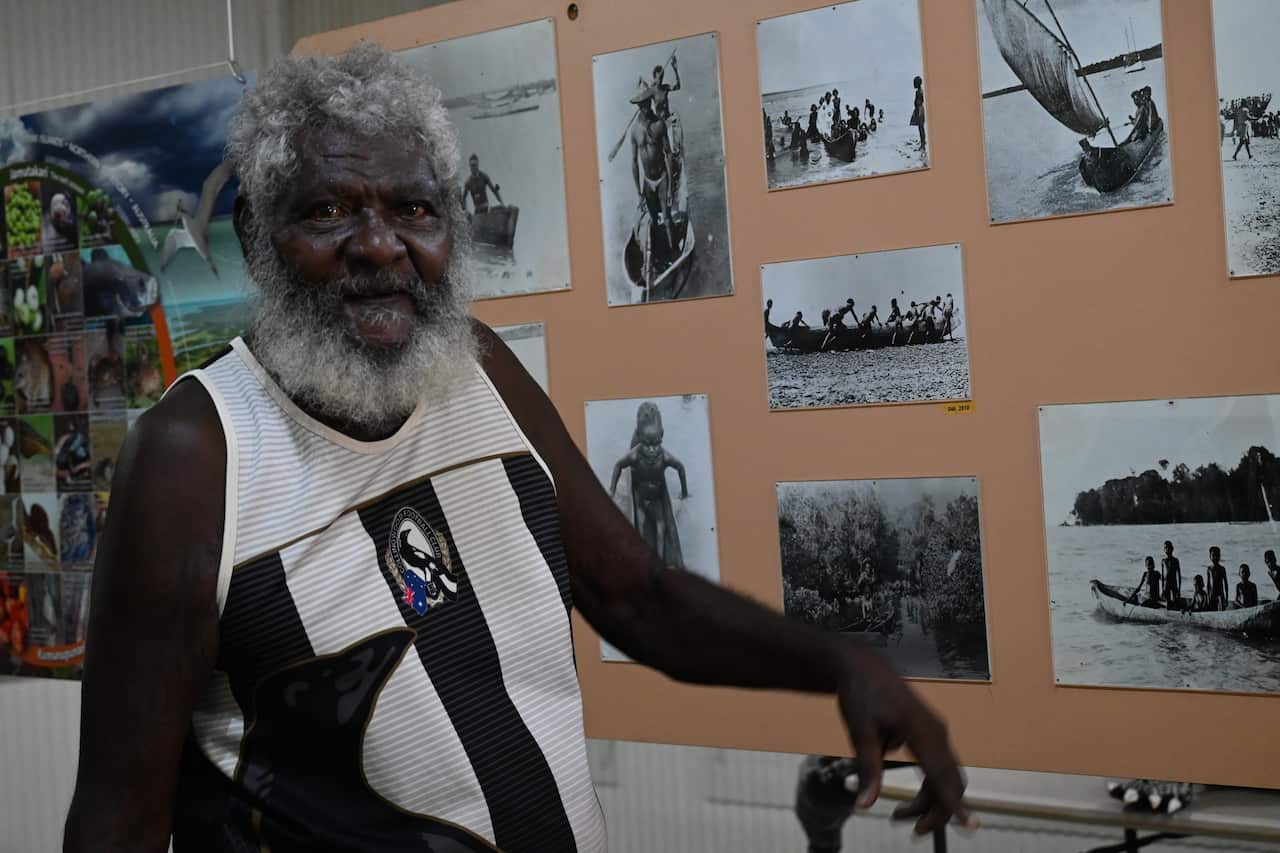 A man in a Collingwood Aussie Rules jumper next to pictures of large dug out canoes on a cork board