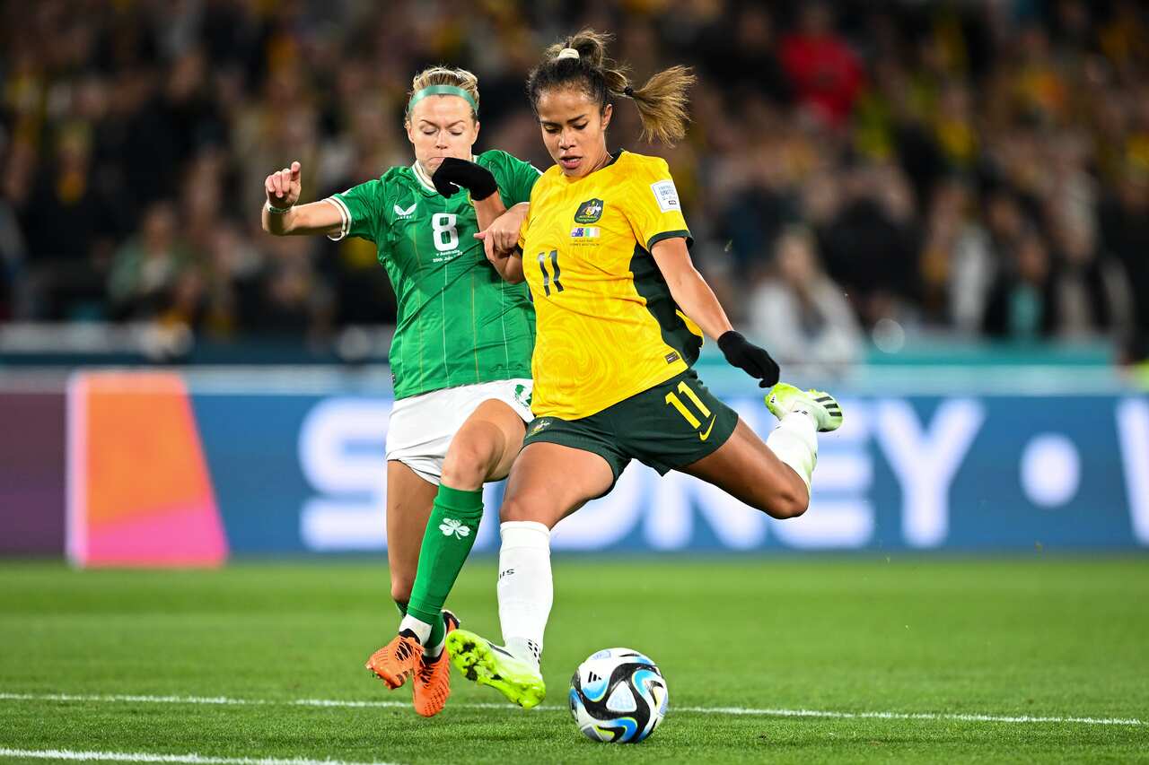 Mary Fowler, wearing a yellow football shirt and green shorts, about to kick the ball