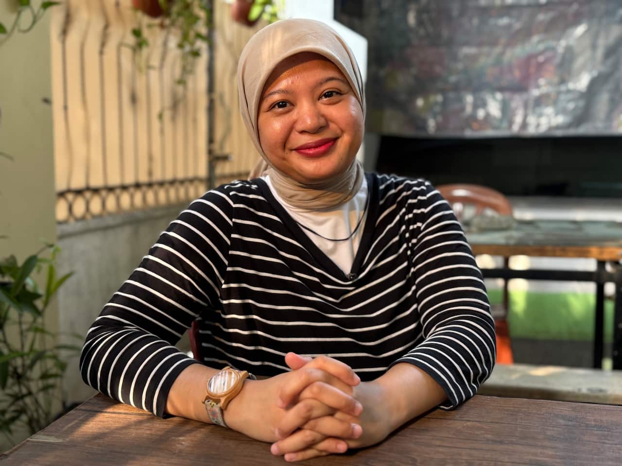 A woman wearing a black-and-white striped top and a head covering smiles as she sits at a table, her hands clasped.