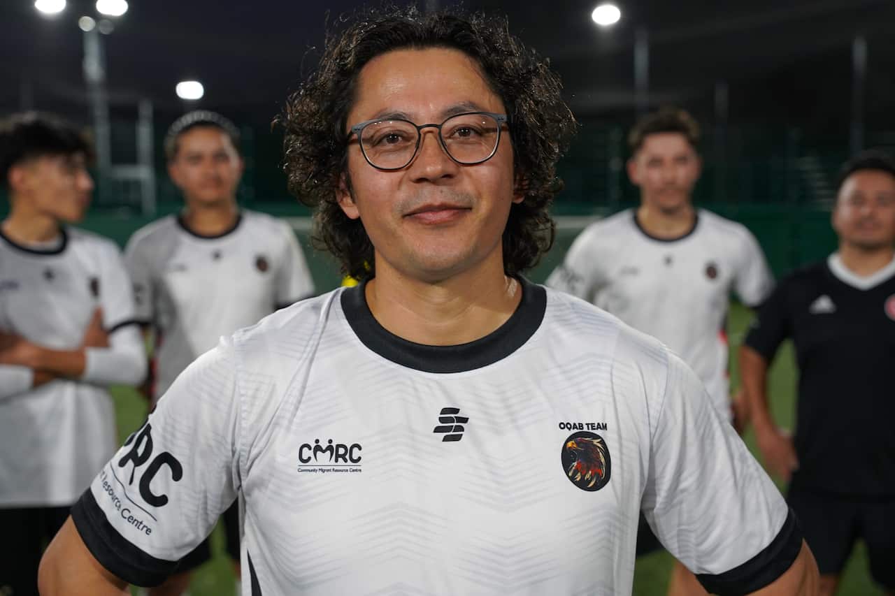 A man standing in a white football shirt with black trim. Other men wearing the same shirt are behind him.