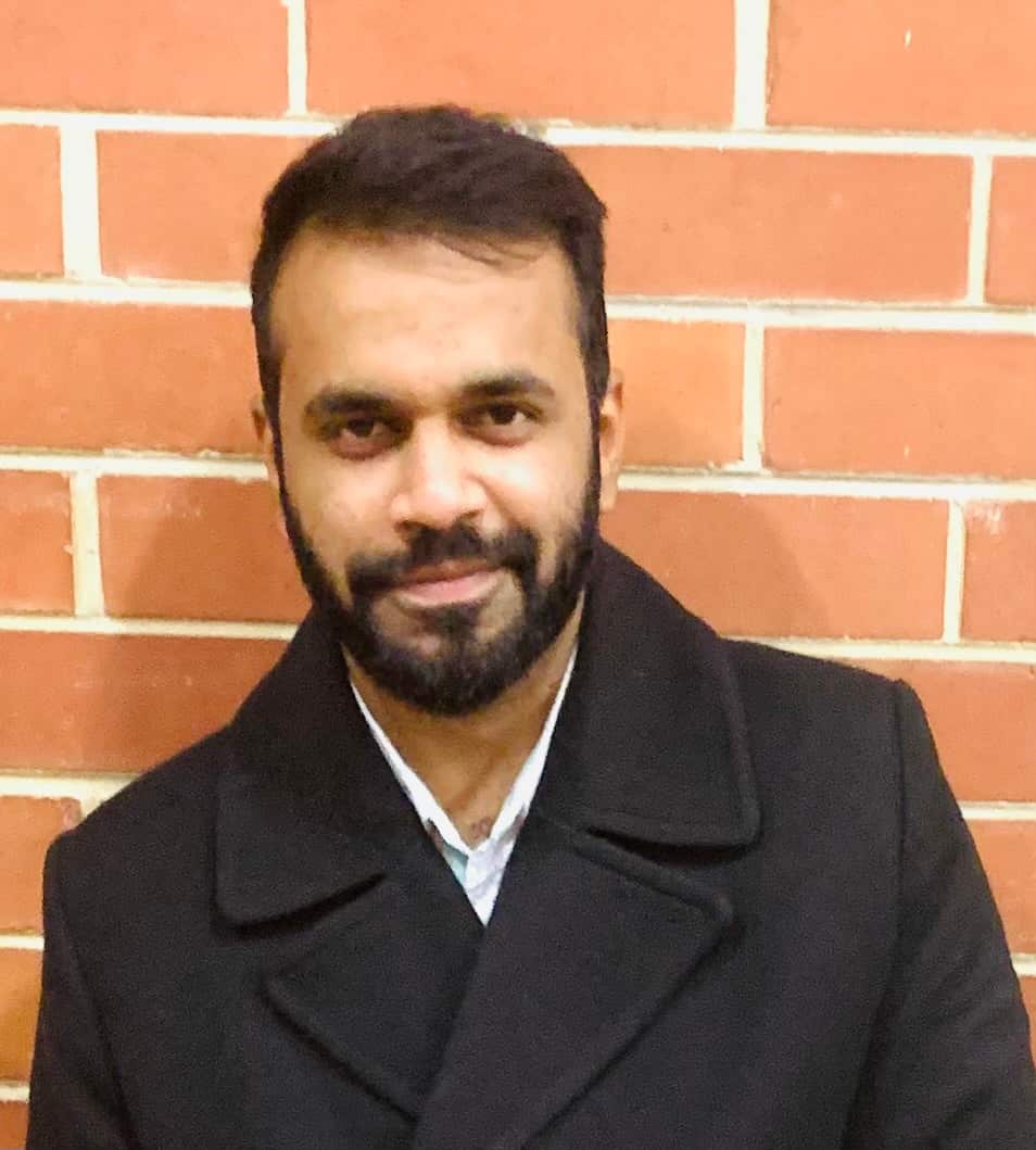 A middle-aged man stands against a brick wall in a white shirt and black coat.