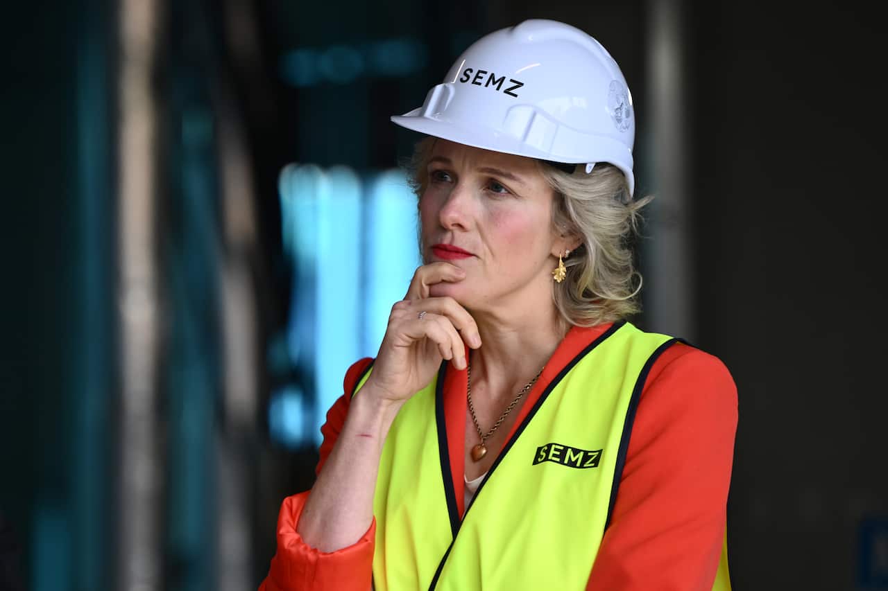 A woman with blonde hair looks contemplative while wearing a hard hat and high-vis vest.
