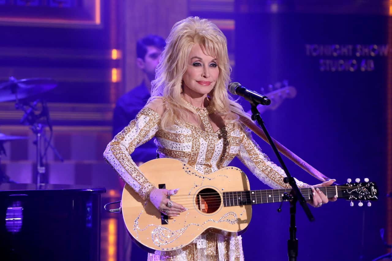 Dolly Parton performing on stage with an acoustic guitar and microphone, wearing a white and gold embellished outfit under purple stage lighting.