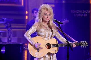 Dolly Parton performing on stage with an acoustic guitar and microphone, wearing a white and gold embellished outfit under purple stage lighting.