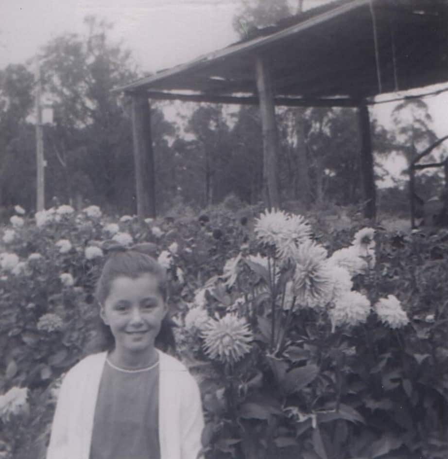 A black and white photo of a young girl standing in a garden with flowers in the background.