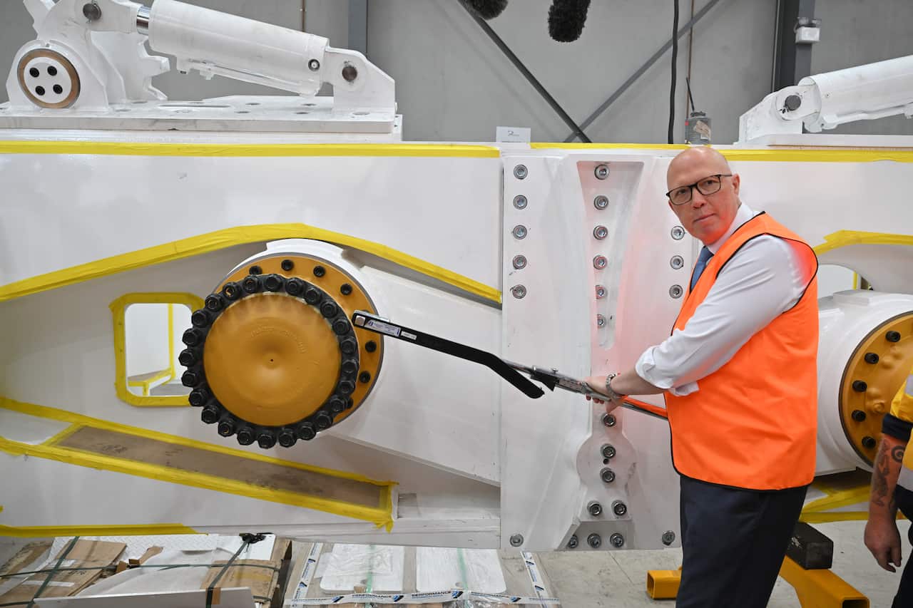 Peter Dutton wearing a high vis vest holds a crank in front of a machine.