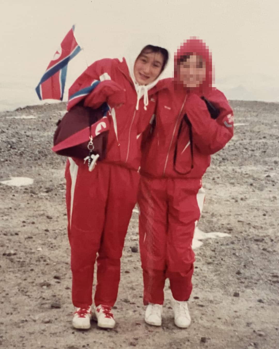 Two teenage girls in red tracksuits. The girl on the left is holding a backpack and a flag of the Democratic People's Republic of Korea