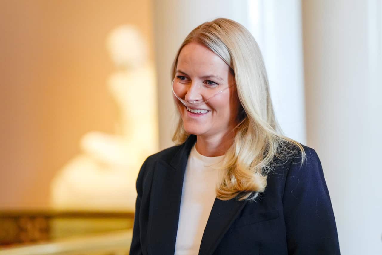 A woman with bright blonde hair looks to one side. She is wearing a suit and has a breathing tube in her nose.