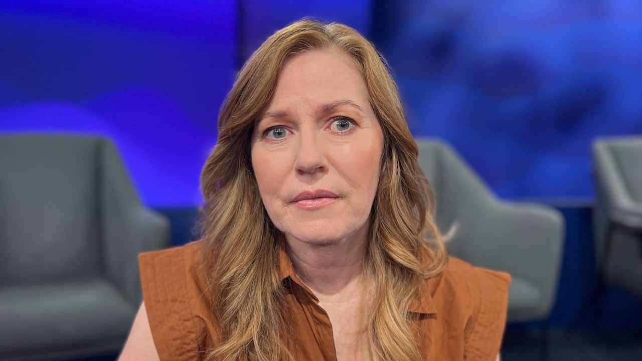 a blonde middle aged woman in a terracotta coloured dress sits in a studio