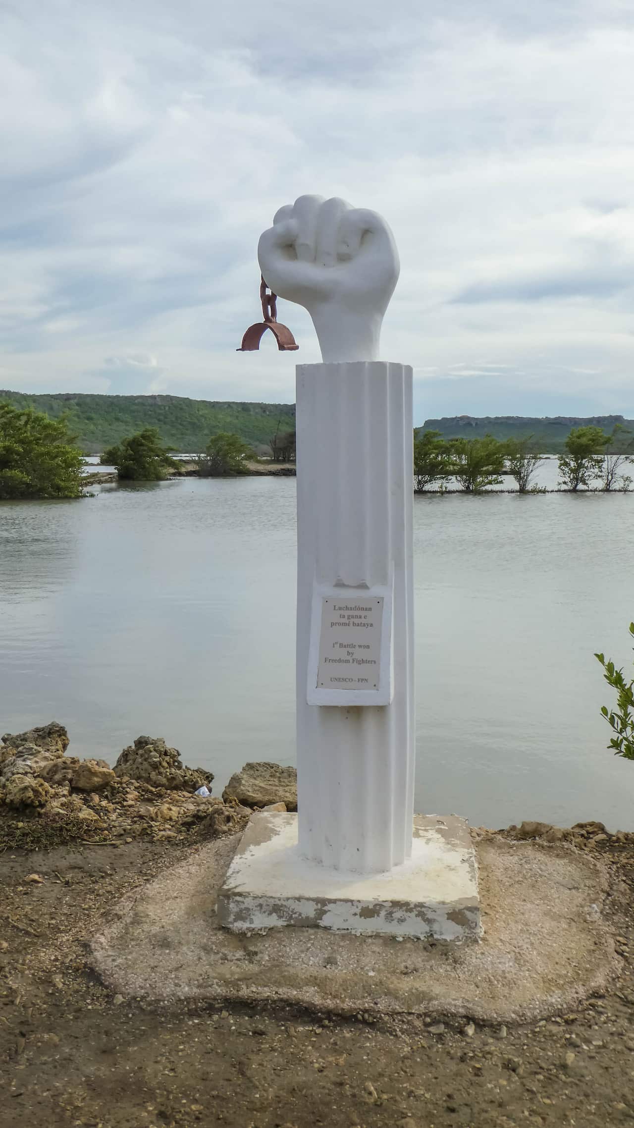 A white statue on a beach, with a raised fist on top of a plinth. The fist is holding a broken manacle. 